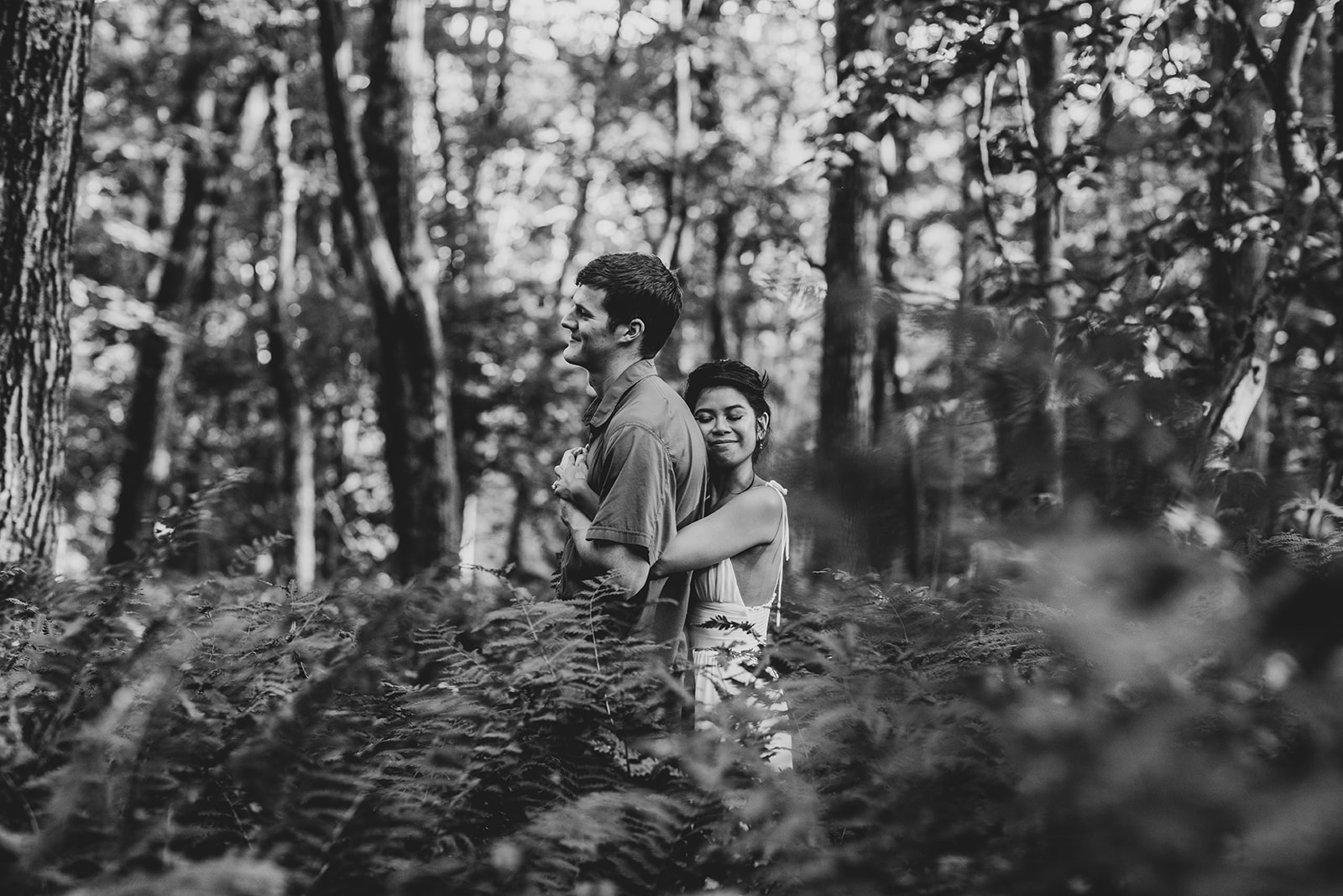 Black and white photo of Jess hugging Joe from behind while standing in tall ferns during their Shenandoah engagement session.