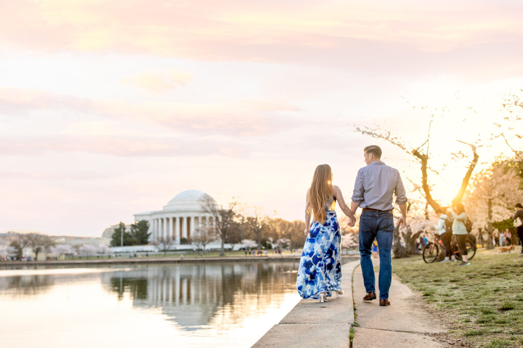 Couple walking on a pathway lined with cherry blossoms at the Tidal Basin in Washington, DC with the Jefferson Memorial in the background at sunrise