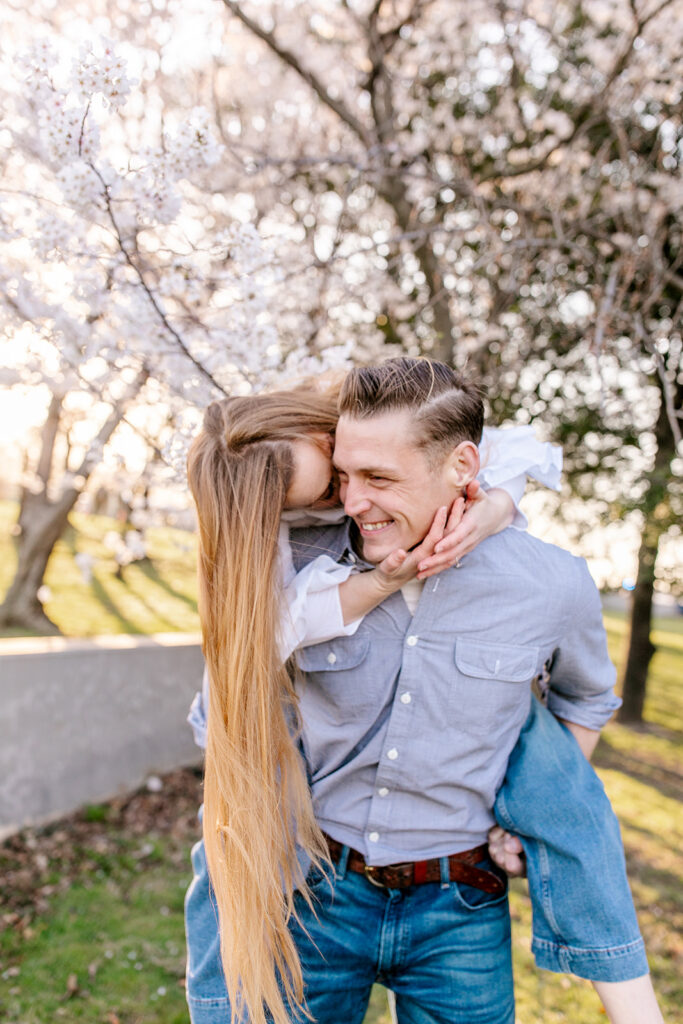 Bride gives groom a kiss during sunrise engagement session in secluded area with cherry blossoms during cherry blossom festival in dc