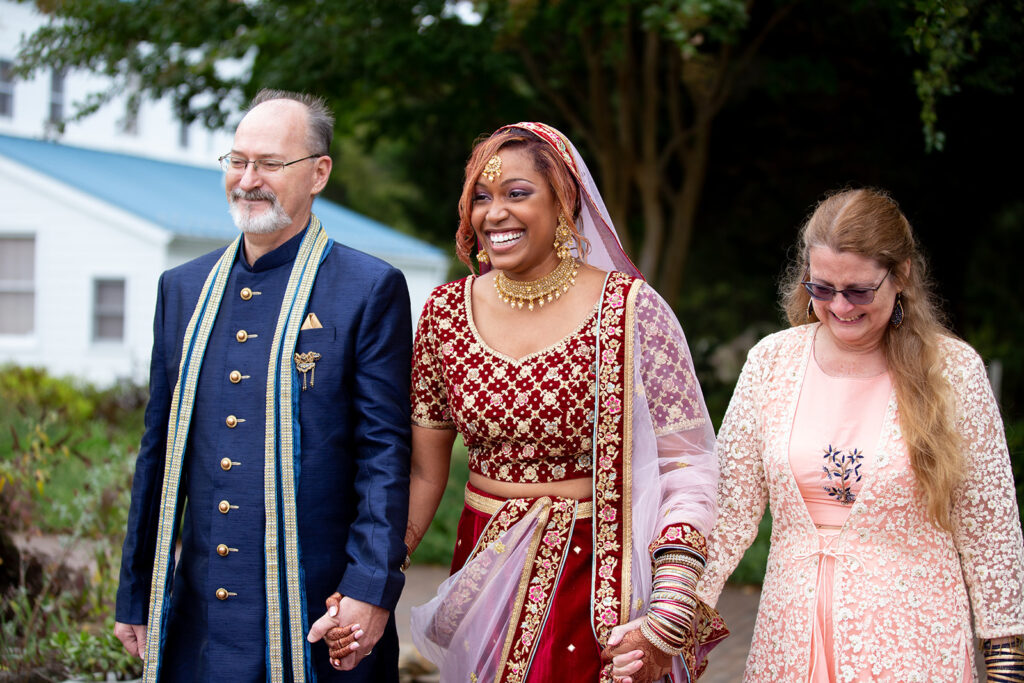 Bride walking down the aisle with her future in-laws during outdoor Indian-American wedding ceremony at The Butterfly Field in Woodford, VA