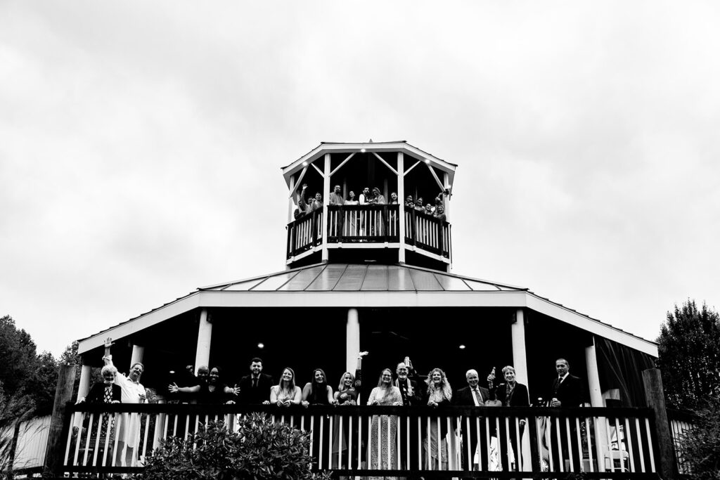 Bridal party posing on rooftop with wedding guests and family waving from below at The Butterfly Field wedding venue in Woodford, VA