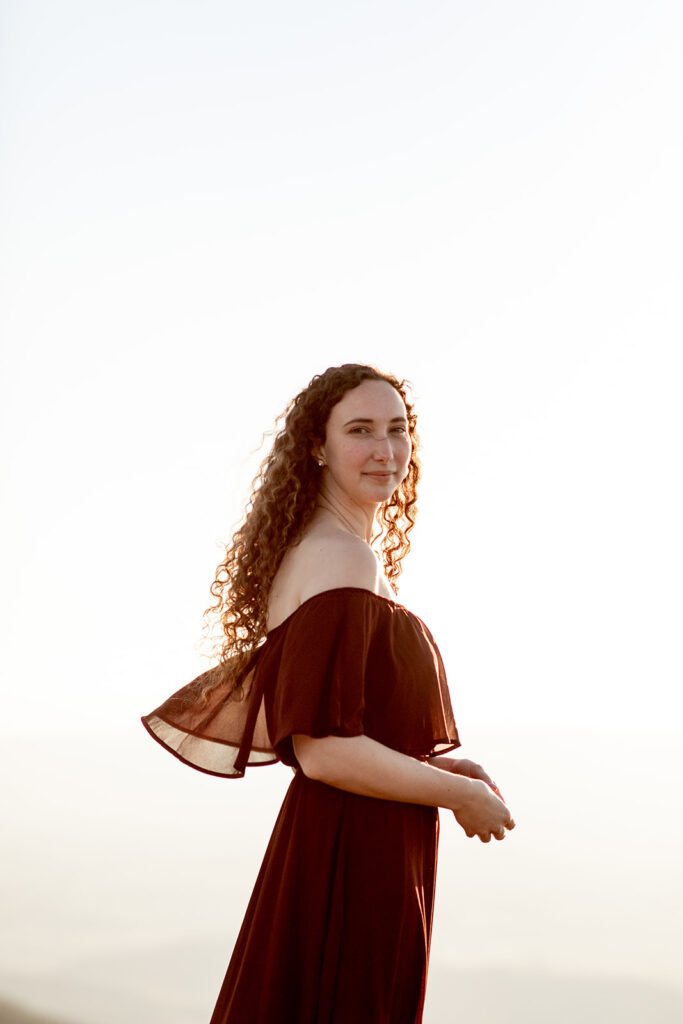 Bride-to-be standing alone on mountaintop in formal burgundy dress during golden hour in Shenandoah Valley
