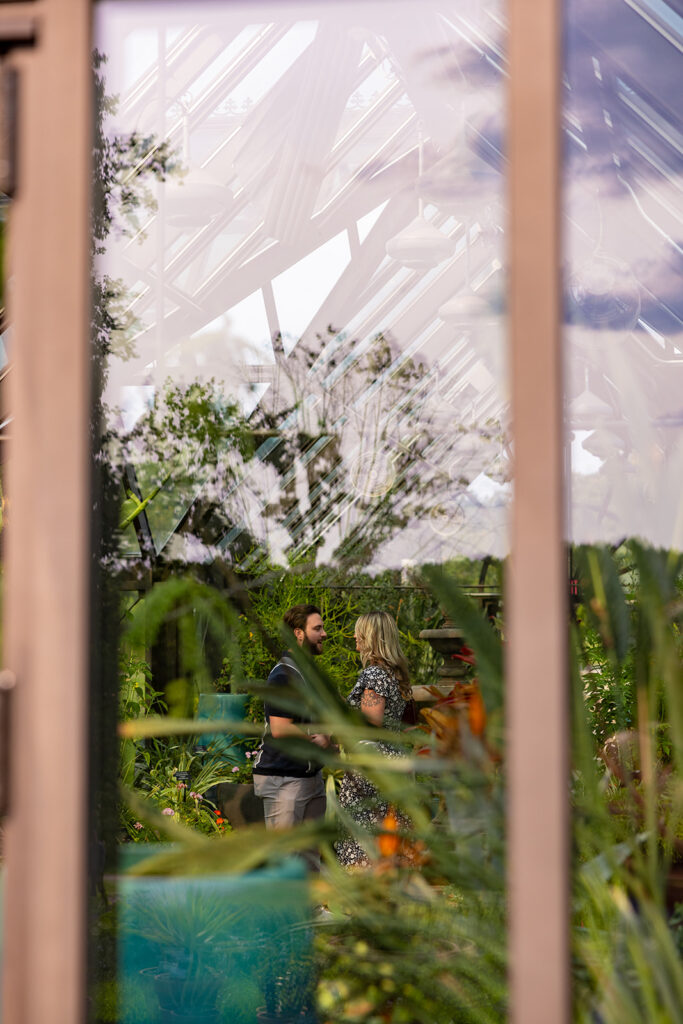 Surprise proposal captured through greenhouse glass at a DC area garden venue, showcasing an intimate engagement moment with a hidden photographer perspective