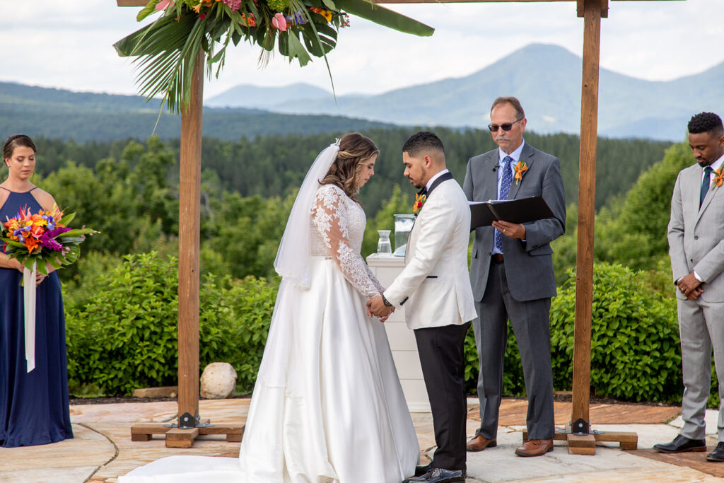 Bride and groom holding hands during a prayer at the altar during their vows with mountain views behind them.