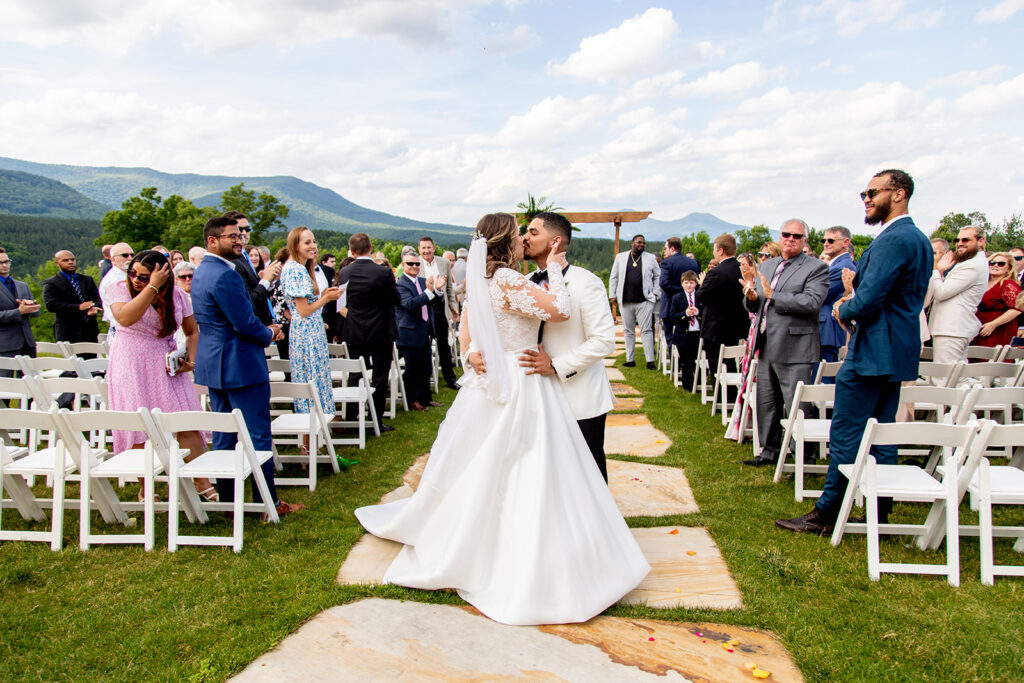 Bride and groom sharing a kiss in the center of the aisle as guests cheer around them during the recessional.