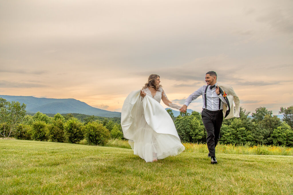 Bride and groom running through a grassy field at The Seclusion, laughing together with mountains in the distance.