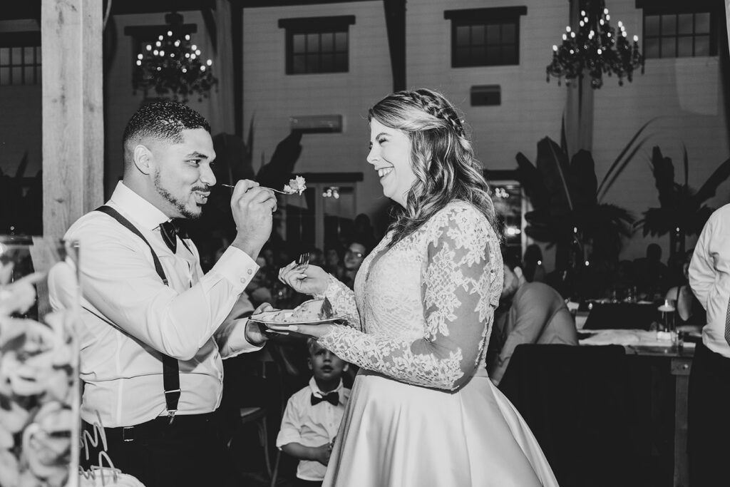 Bride and groom feeding each other cake with guests watching, black-and-white photo.