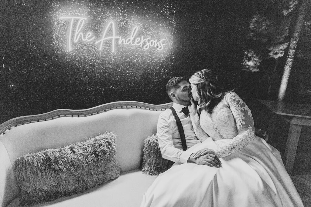 Bride and groom sitting together on a white sofa under a neon “The Andersons” sign.