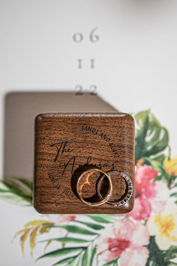 Wedding rings displayed on a personalized wooden ring box over the tropical invitation suite.