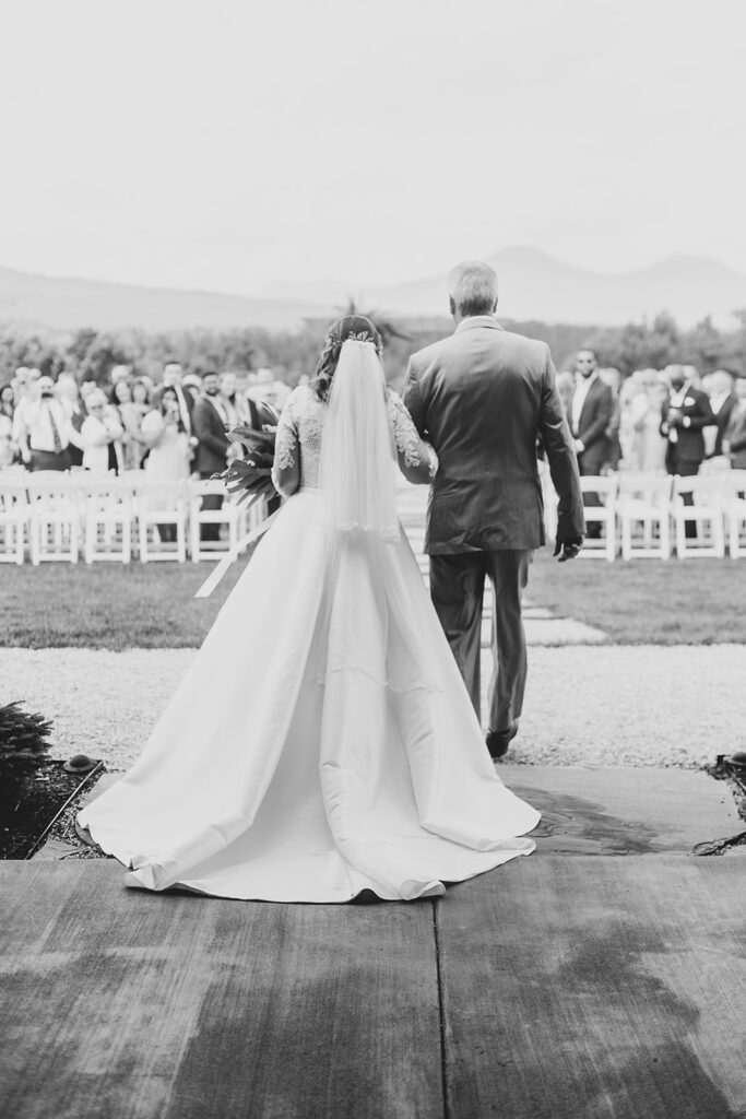 Bride walking down the aisle with her father toward the mountain-view ceremony space, black-and-white photo.