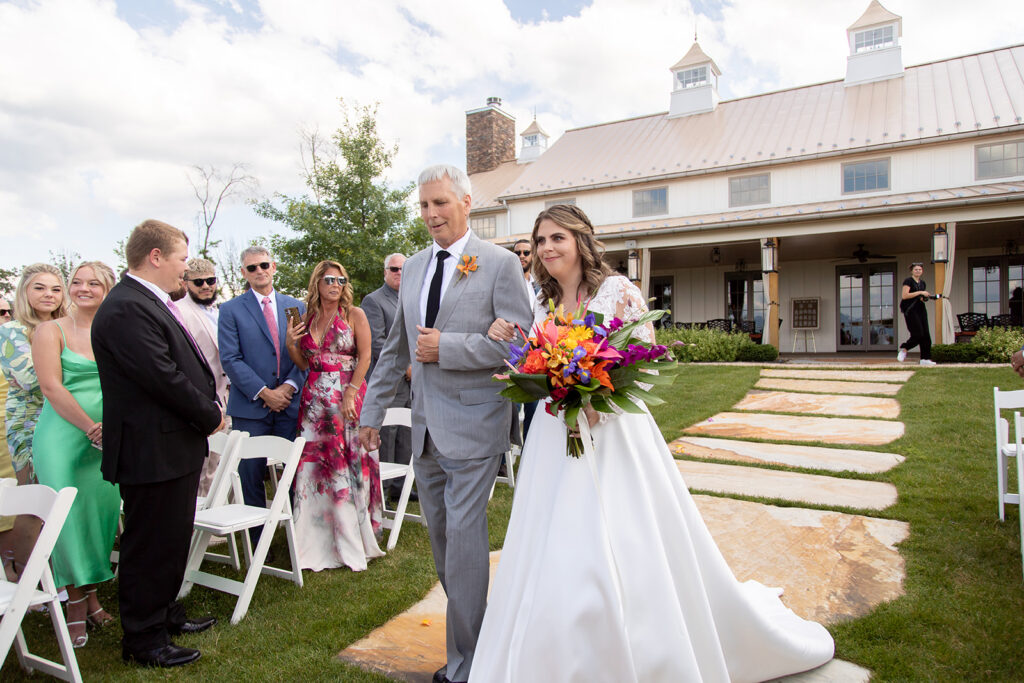Bride walking down the aisle holding a colorful tropical bouquet, escorted by her father.