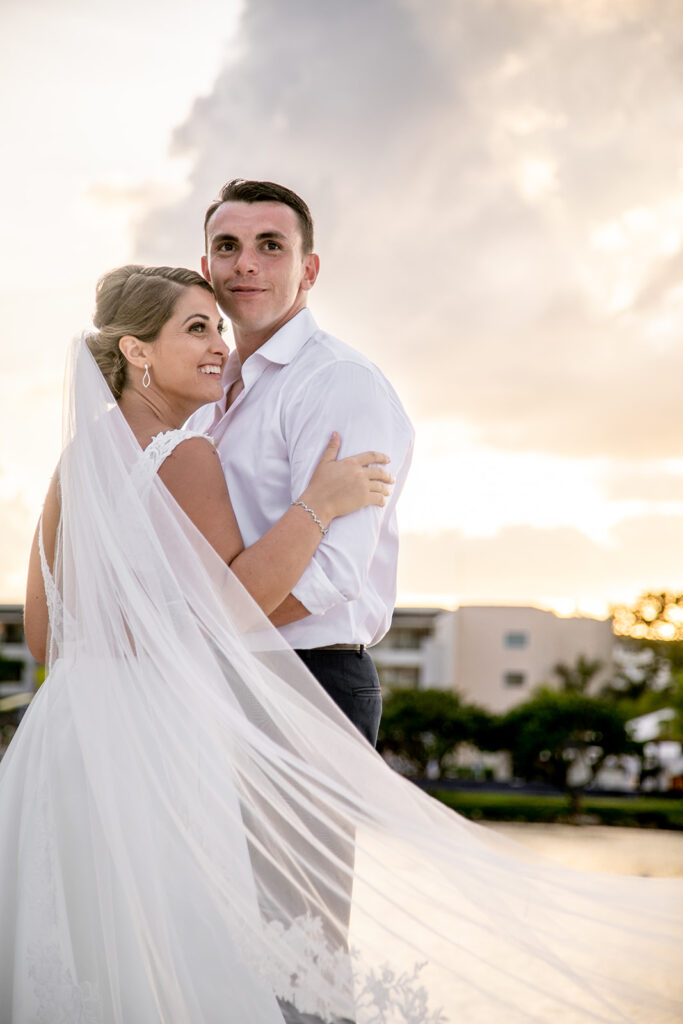Bride and groom standing together on the beach at golden hour, veil blowing in the breeze.