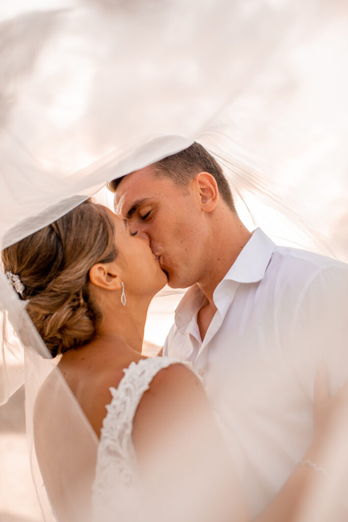 Bride and groom kissing under the bride’s veil with soft sunset light filtering through.