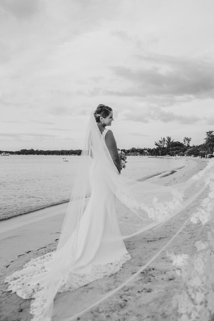 Bride standing alone on the beach with her veil trailing behind her in black and white.