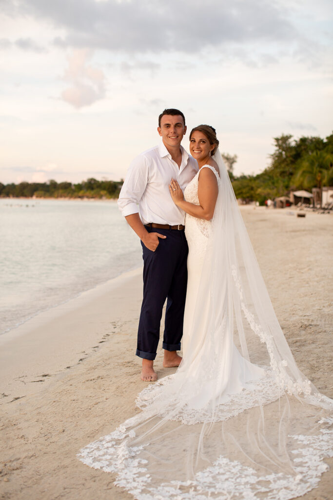 Bride and groom standing barefoot on the beach at sunset with the bride’s long veil trailing behind her.