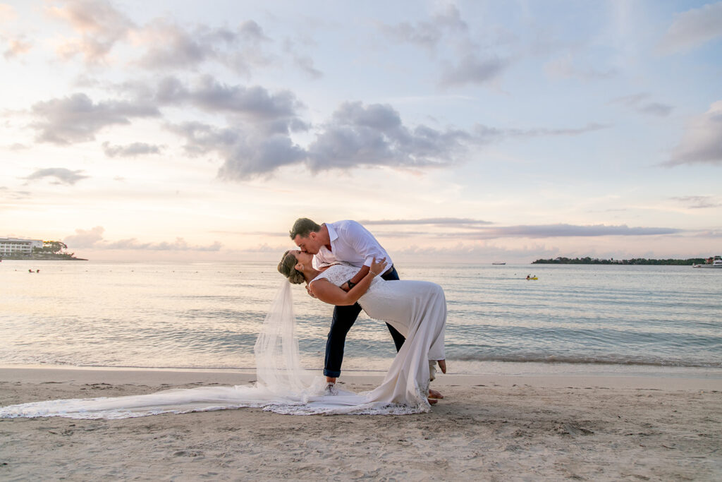 Groom dipping the bride in the sand at sunset with pastel skies behind them.