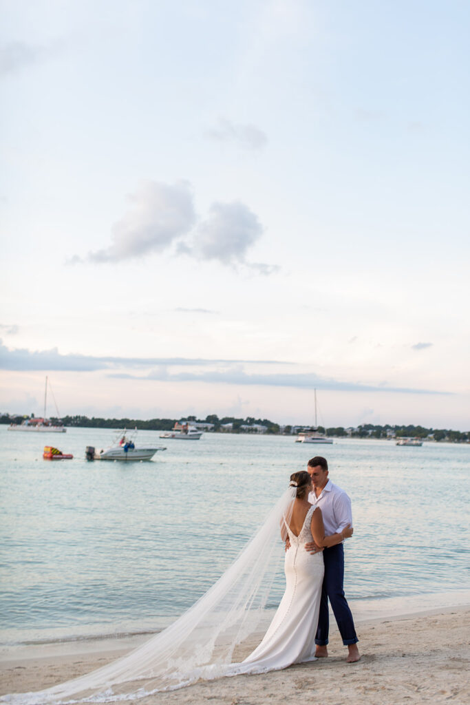 Couple standing in the shallow water at dusk with sailboats in the distance.