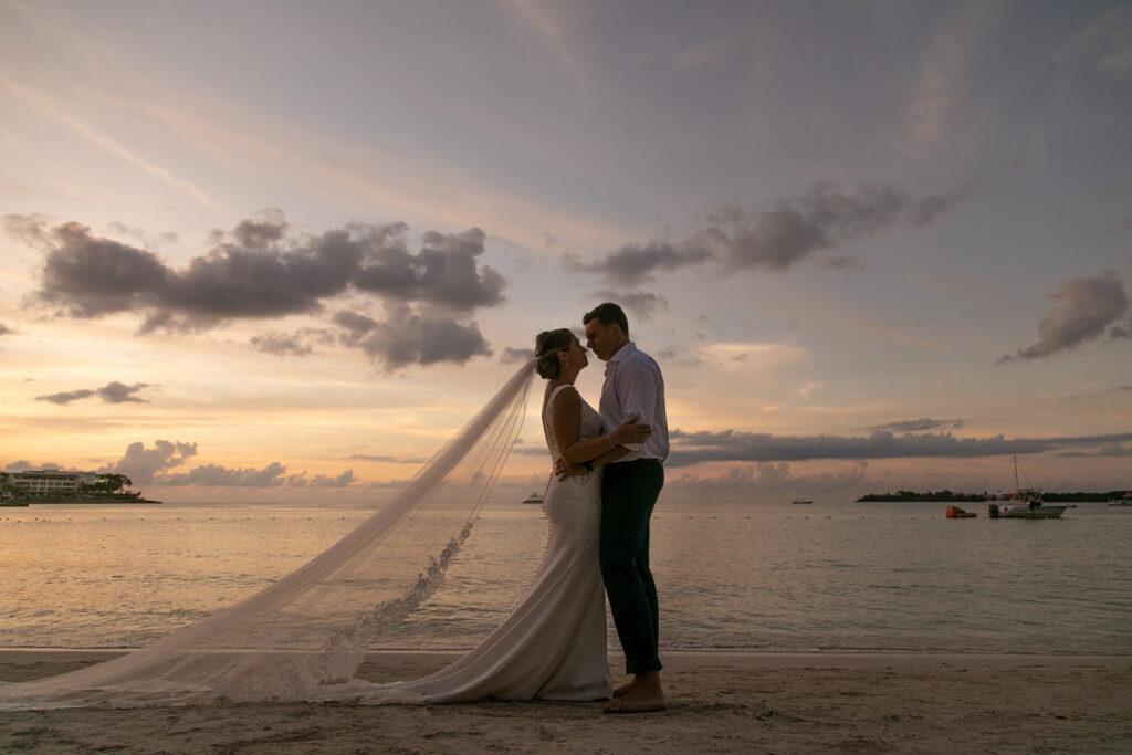 Bride and groom standing together during blue hour with the veil cascading across the sand as the sky glows behind them.