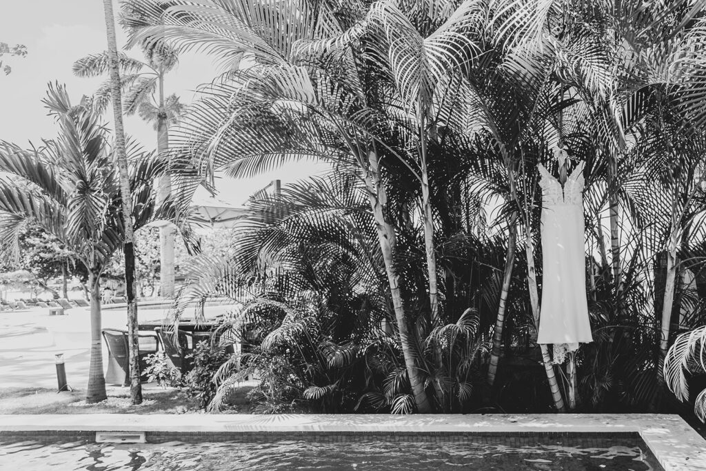 Black and white photo of the bride’s wedding gown hanging from palm trees beside the pool at Royalton Negril Resort.