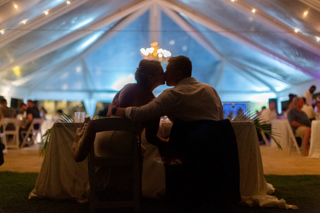 Bride and groom sitting at their sweetheart table, silhouetted as they kiss under glowing lights.