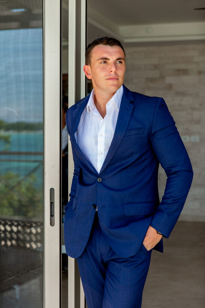 Groom standing confidently in a navy suit at the sliding doors of his resort room.