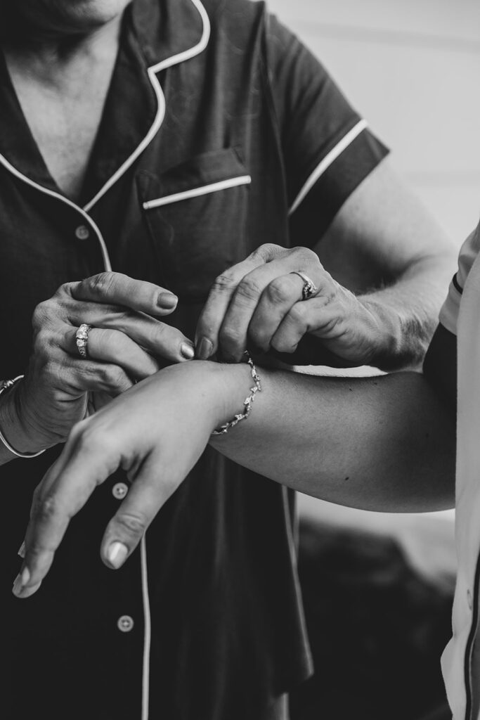 Black and white image of the bride’s mother fastening a bracelet onto the bride’s wrist.