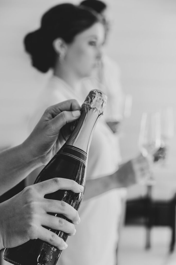 Black and white image of champagne being opened while the bride holds a glass during getting-ready preparations.