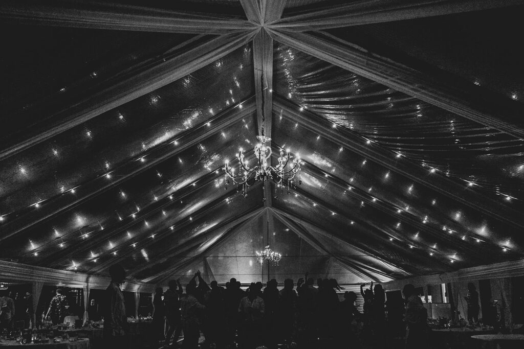 Silhouette of guests dancing beneath a clear reception tent glowing with chandelier and string lights.