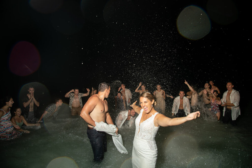 Bride and groom laughing in the ocean while friends celebrate behind them during their destination wedding afterparty.