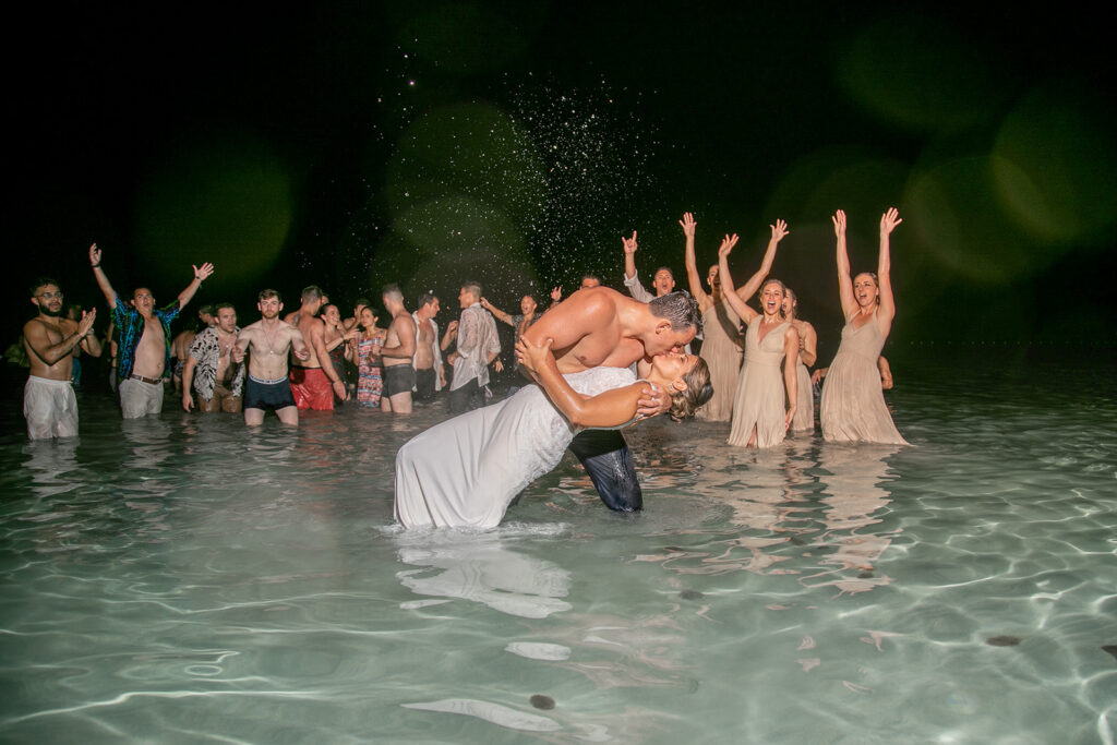 Groom dips the bride for a dramatic kiss in the ocean at night, surrounded by cheering wedding guests during their Jamaica wedding celebration.