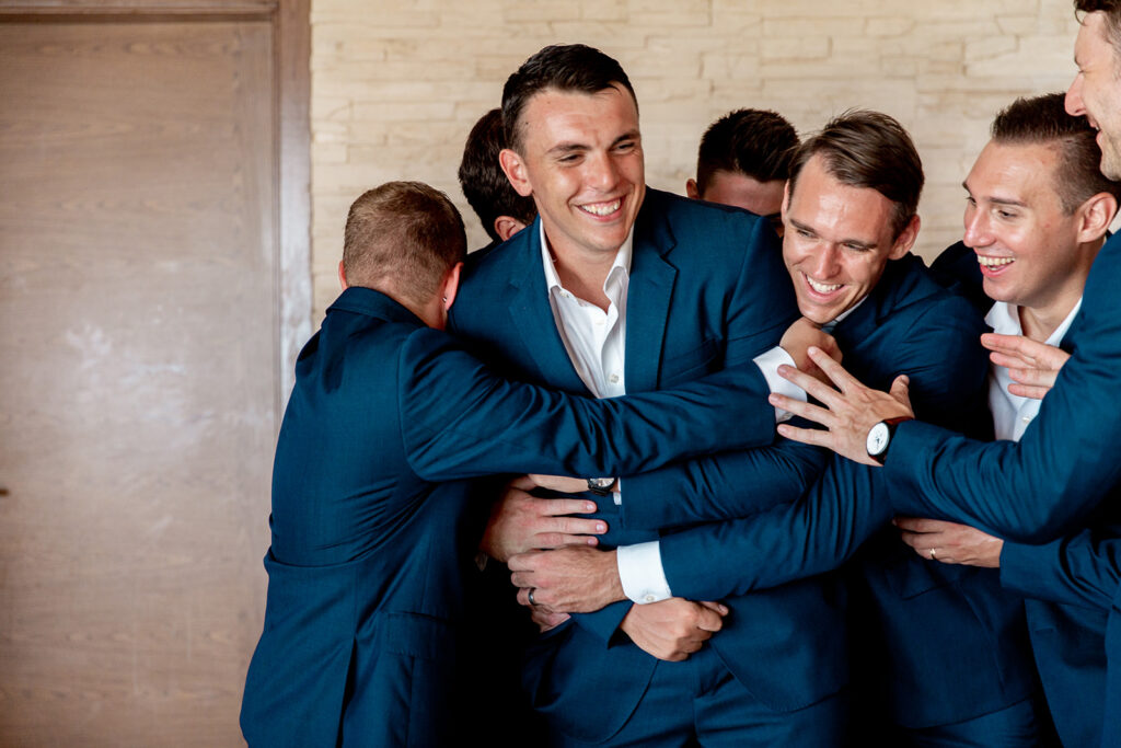 Groomsmen wrapping the groom in a big group hug, all laughing and celebrating.