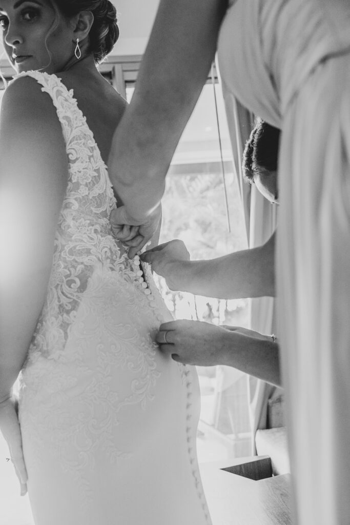 Black and white close-up of bridesmaids buttoning the back of the bride’s lace wedding gown.