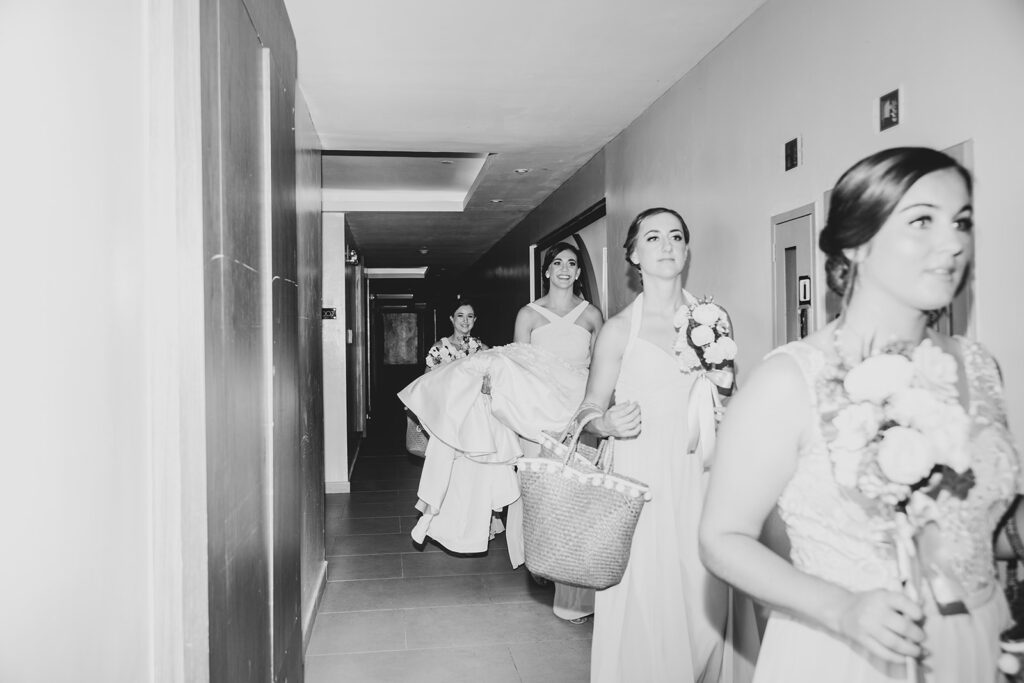 Black and white photo of bridesmaids walking down the resort hallway carrying bouquets and the bride’s dress.