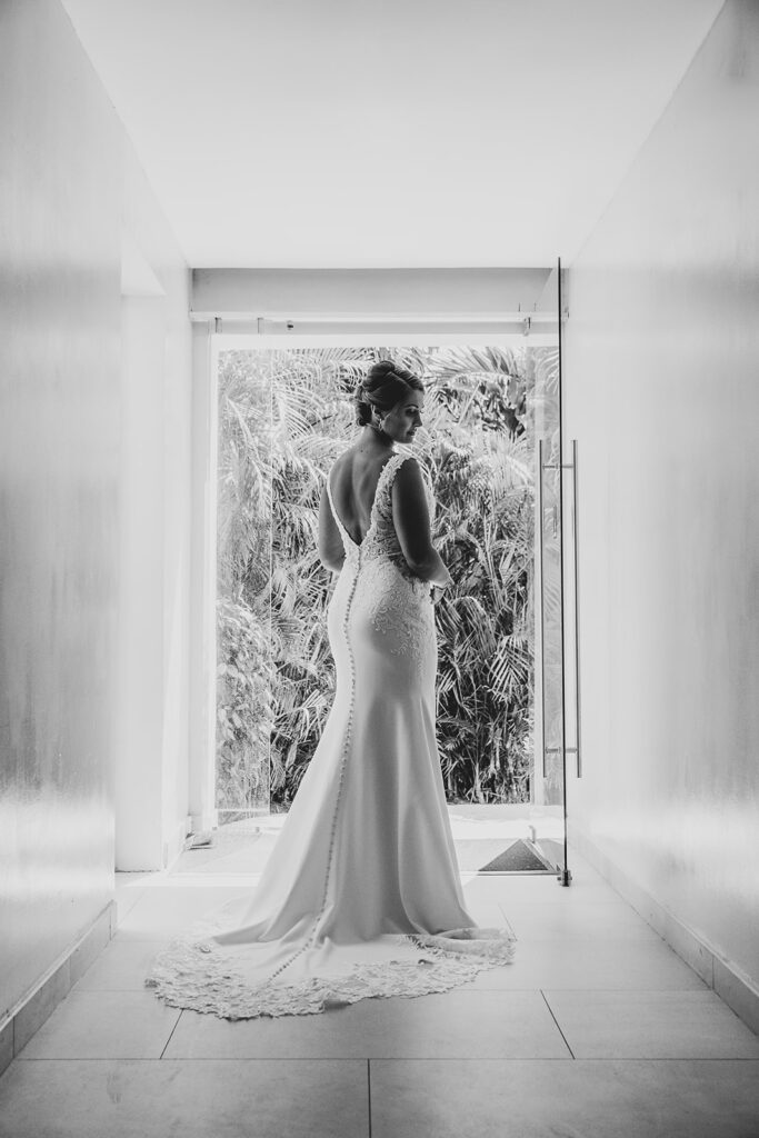 Black and white portrait of the bride standing in a bright hallway, looking out toward the greenery in her fitted gown.
