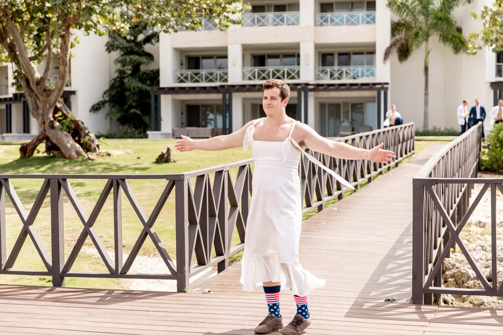 Best man standing on the dock with his arms open, dressed as a fake bride, smiling as he prepares for the faux first look.