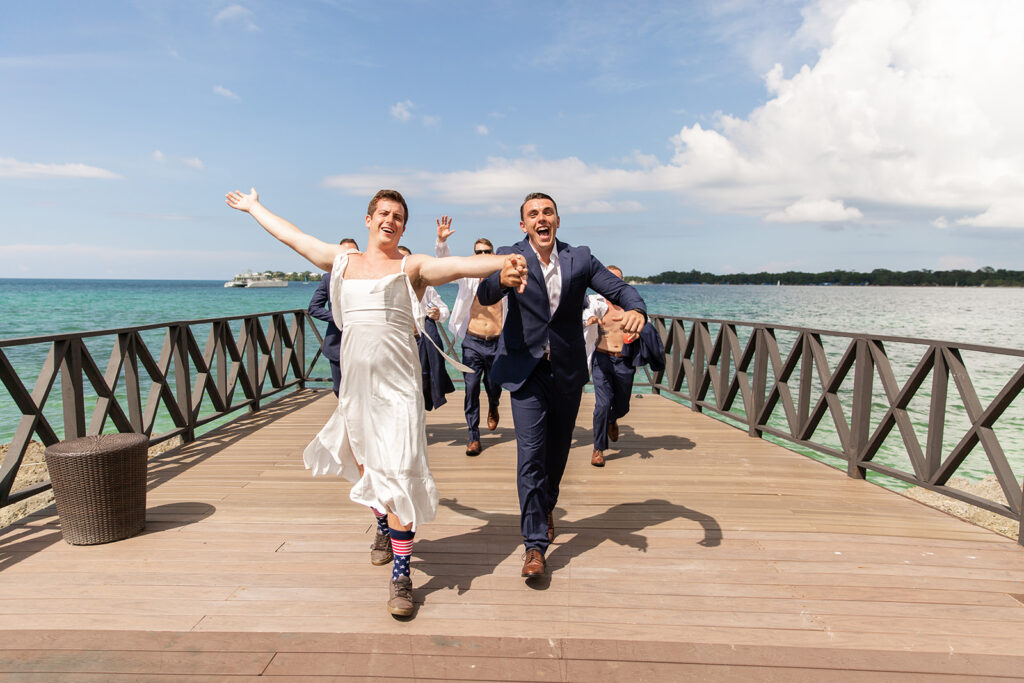 Groom and friend dressed in a wedding gown running joyfully down the dock together with the ocean behind them.