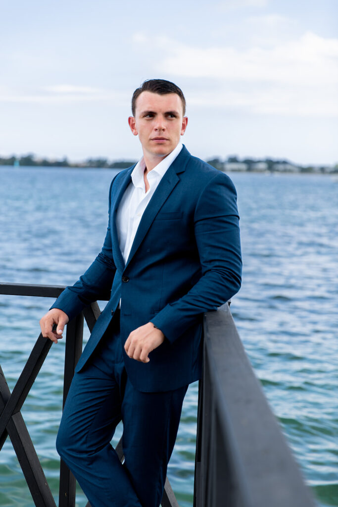 Groom standing alone against a wooden railing with blue ocean water behind him.