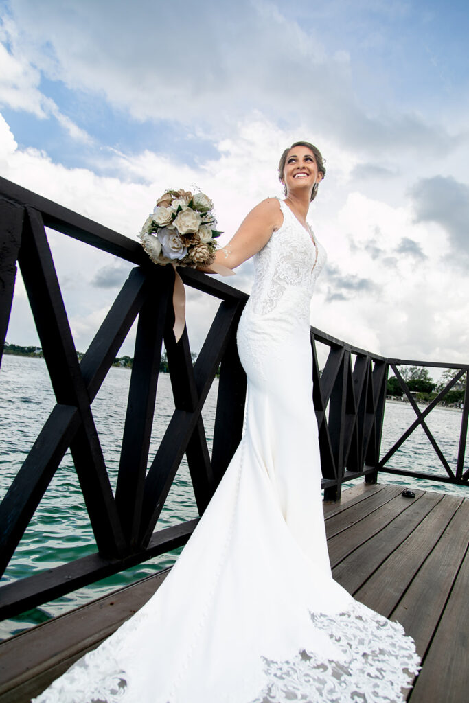 Bride leaning against a wooden railing holding her bouquet with the ocean behind her.