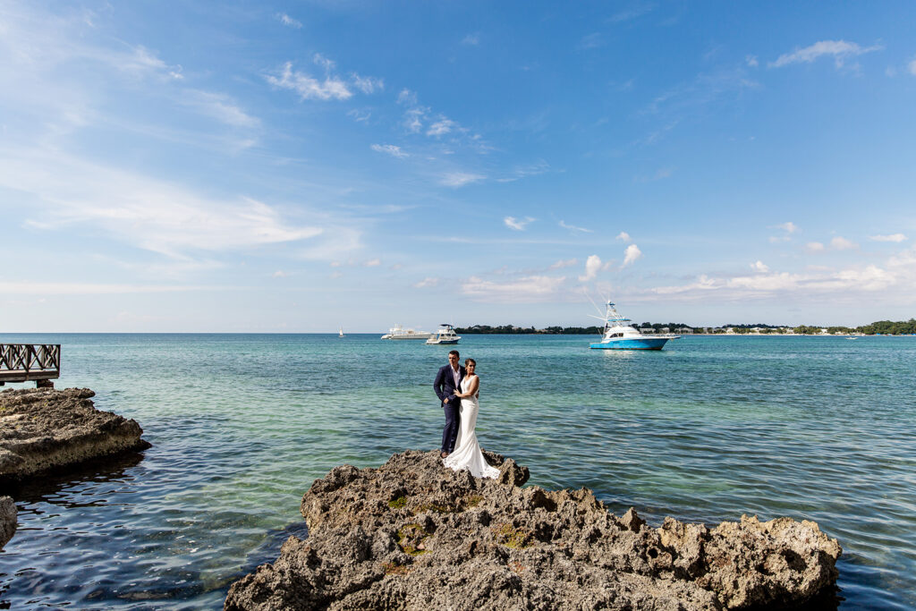 Bride and groom standing on jagged ocean rocks at Royalton Negril with turquoise water behind them.