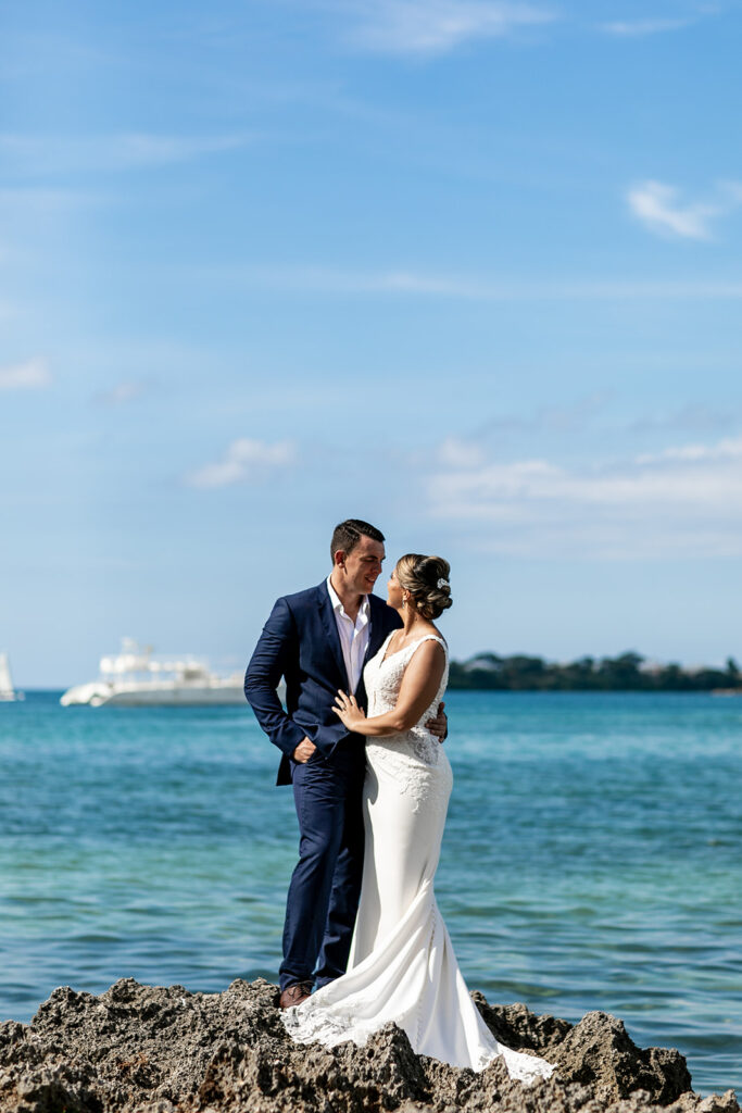 Bride and groom standing on a rock formation, leaning into each other with the ocean in the background.