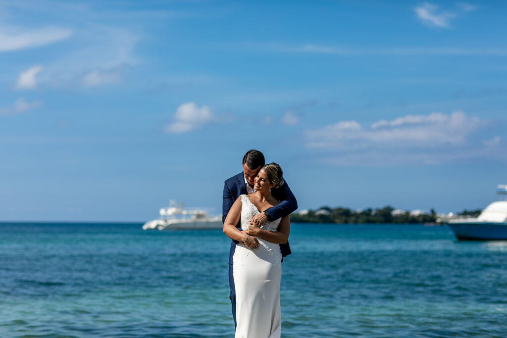 Groom hugging bride from behind as they smile with calm Caribbean water behind them.