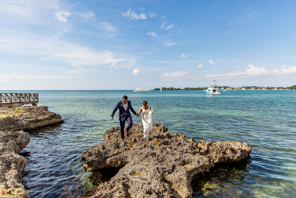 Bride and groom walking hand-in-hand across textured ocean rocks with clear blue water below.