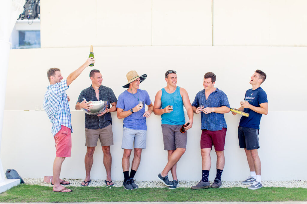 Full group photo of the groom and groomsmen lined up against a white wall, holding cigars and drinks.