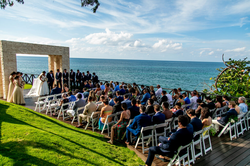 Wide view of the oceanfront ceremony with guests seated facing the stone arch.