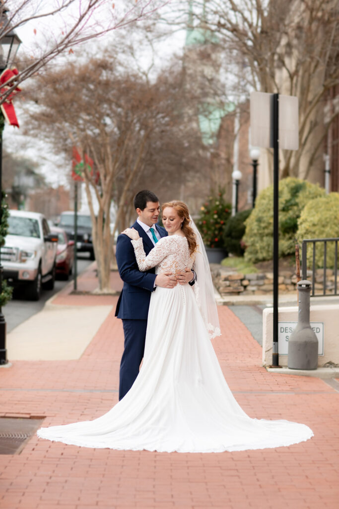 Bride and groom standing on a brick sidewalk in downtown Fredericksburg, with the bride’s long train flowing behind her.