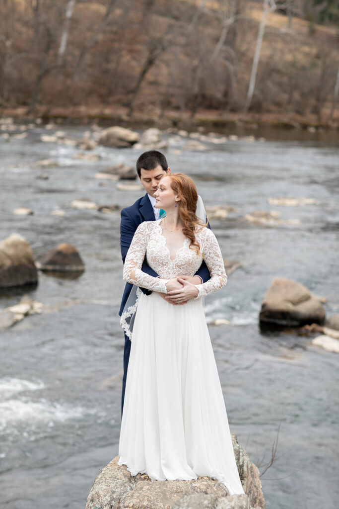 Bride and groom standing on a rock in the river, with the groom embracing the bride from behind.