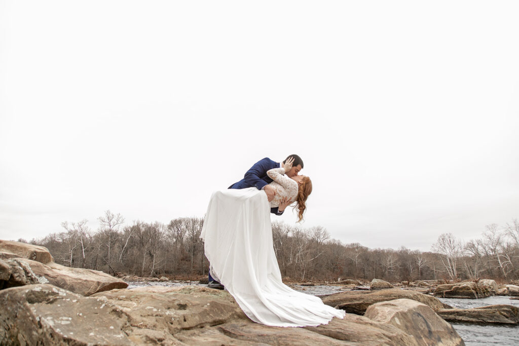 Groom dipping the bride backward for a kiss on a large river rock, with her long train sweeping across the stone.