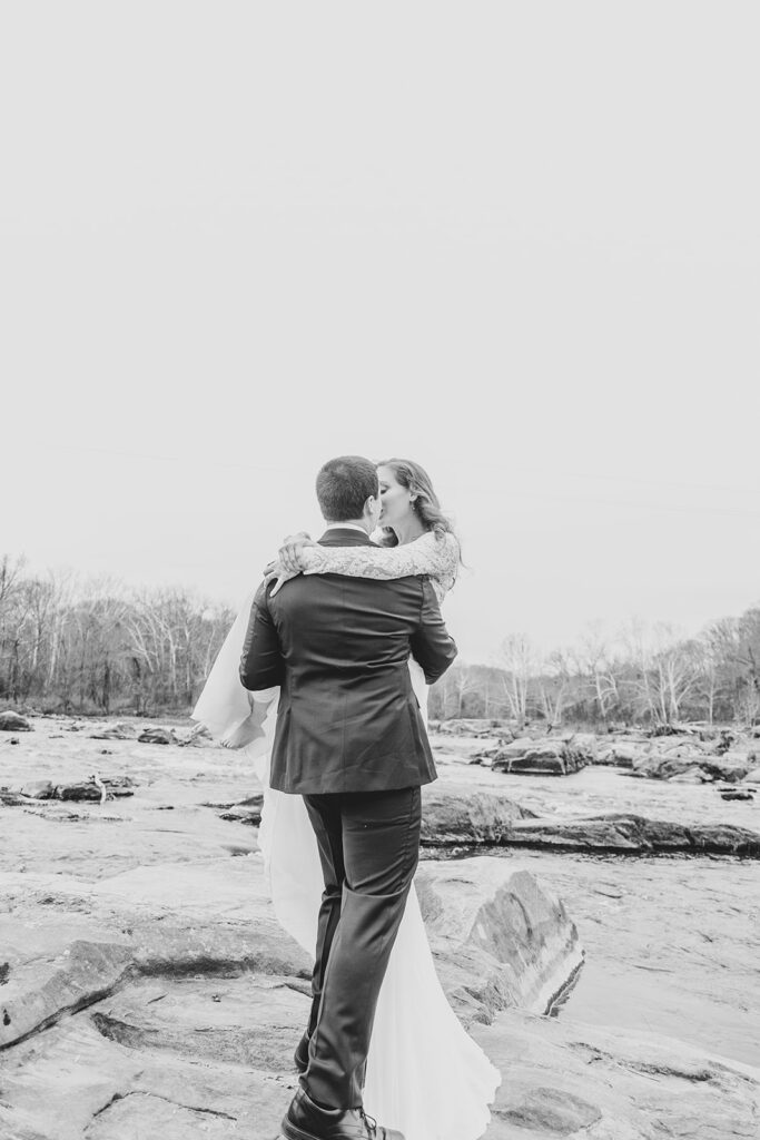 Groom carrying the bride across a rocky riverbank, black-and-white portrait.