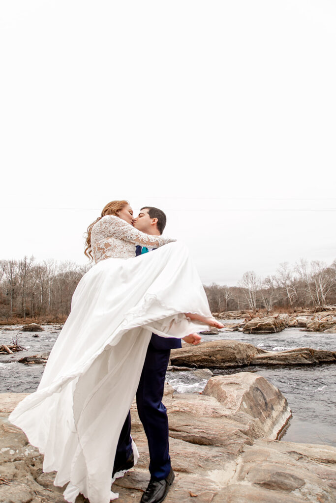 Groom lifting the bride in his arms on a rock beside the river, her dress billowing in the wind.