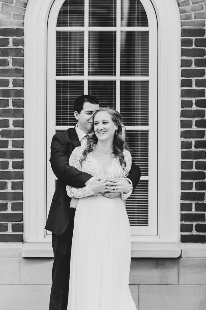 Bride laughing as the groom hugs her from behind in a black-and-white portrait by the courthouse window.